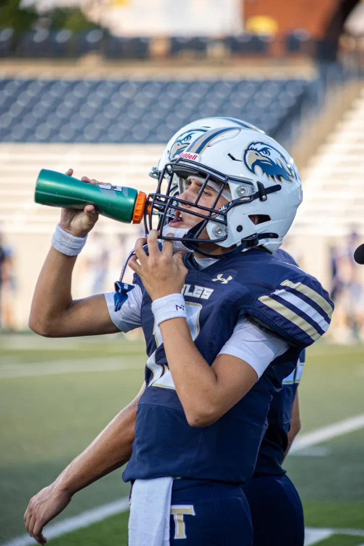 A helmeted football player drinks from a water bottle on the sideline.