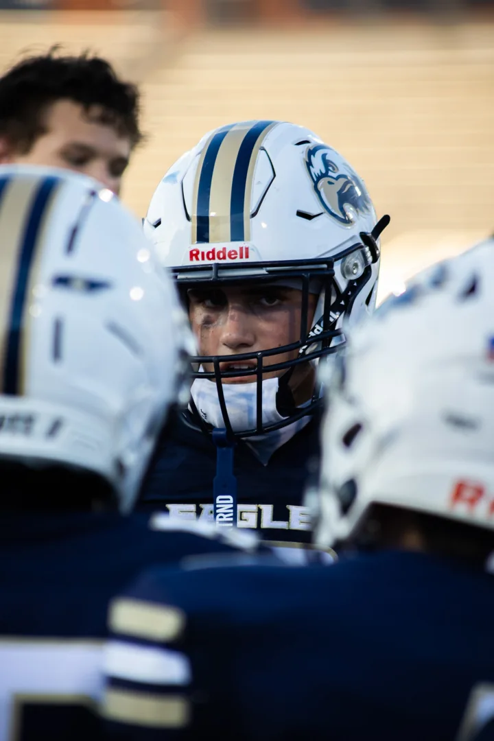 A helmeted football player looks forward from the middle of a team huddle.