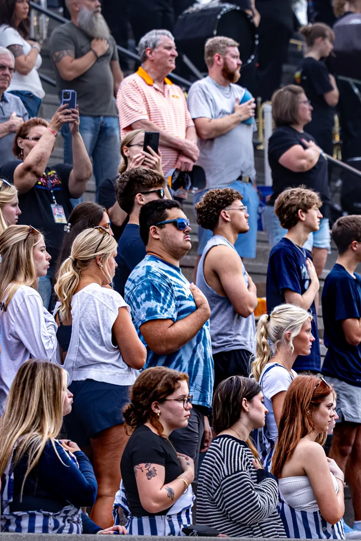 Spectators stand and watch from a crowded football grandstand.