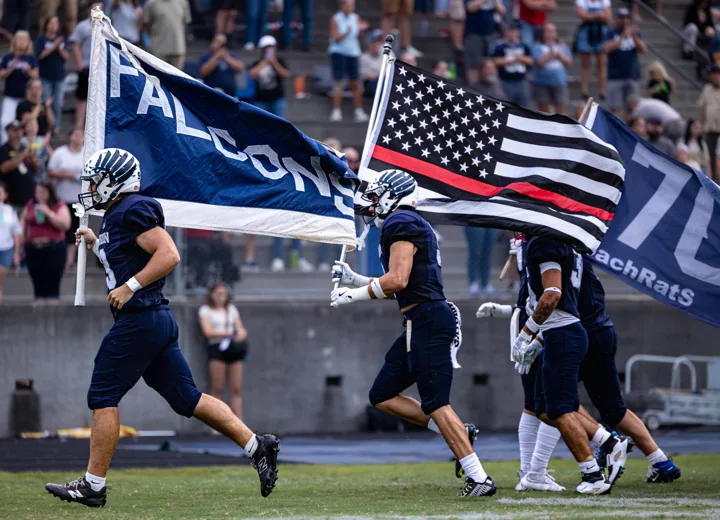 Football players run onto the field carrying team and United States flags.