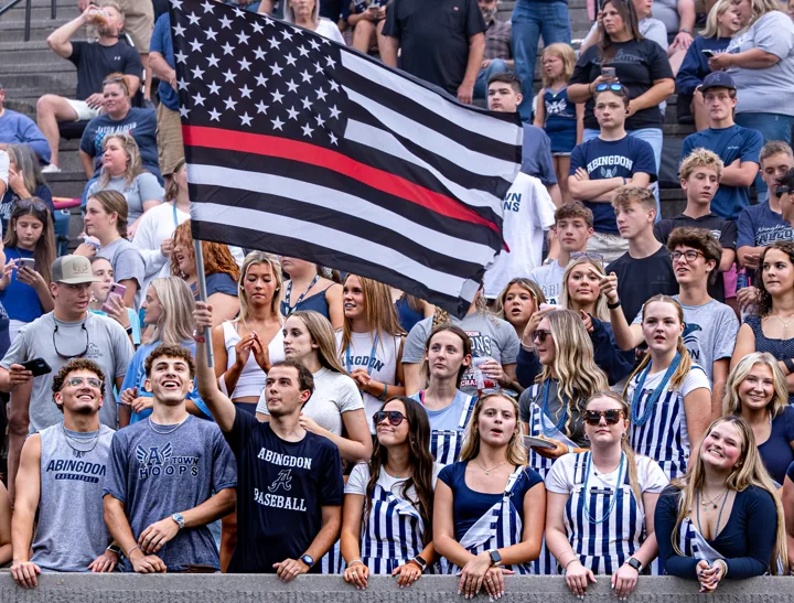 A crowd cheers beneath a large United States flag at a football game.