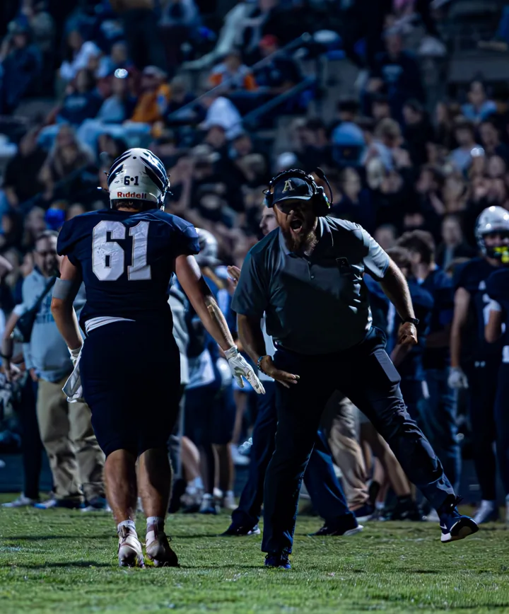 A football coach speaks with a player on the sideline during a night game.