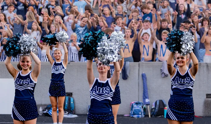 Cheerleaders raise silver pom-poms in front of a crowded football grandstand.