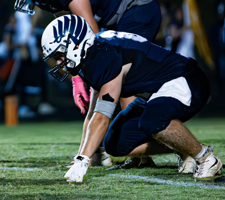 A helmeted football player crouches low with one hand on the turf.