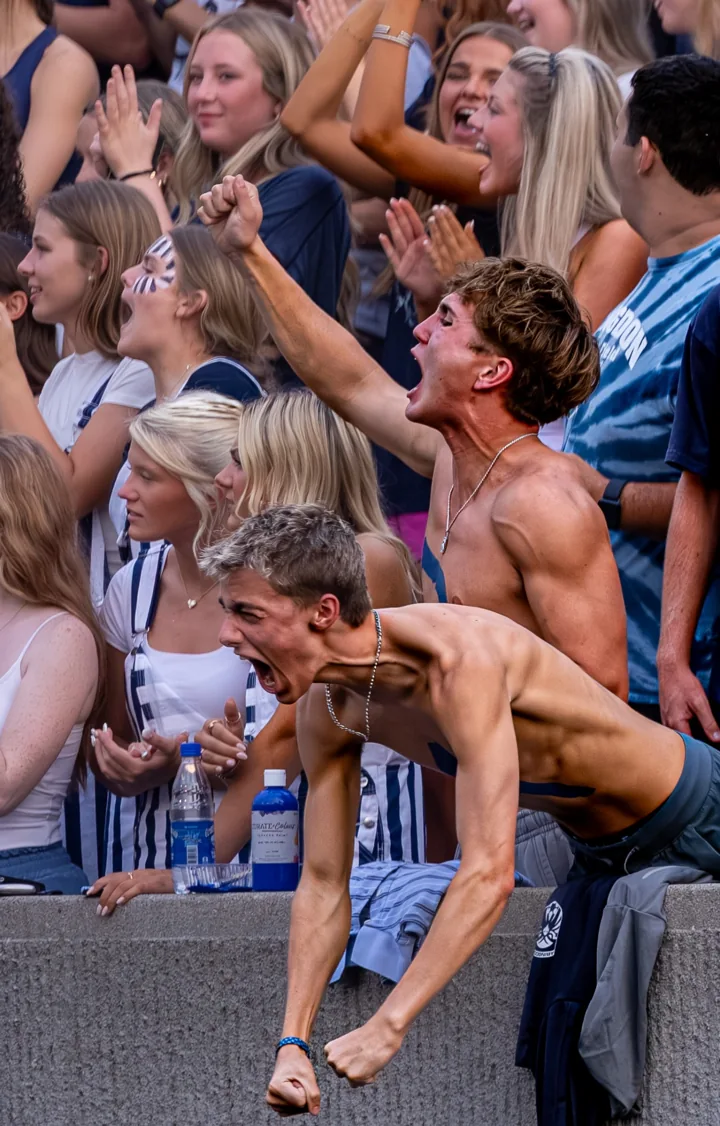 Students lean over the front of the grandstand and cheer toward the field.