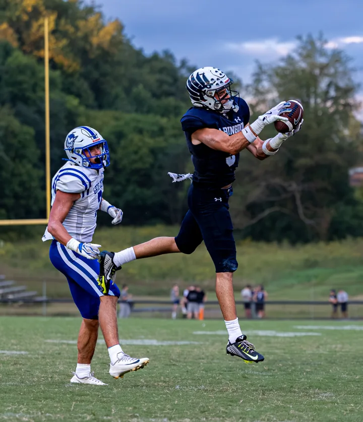 A football receiver leaps to catch the ball as a defender follows behind.