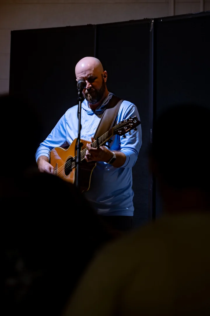 A musician sings into a microphone while playing an acoustic guitar under a spotlight.