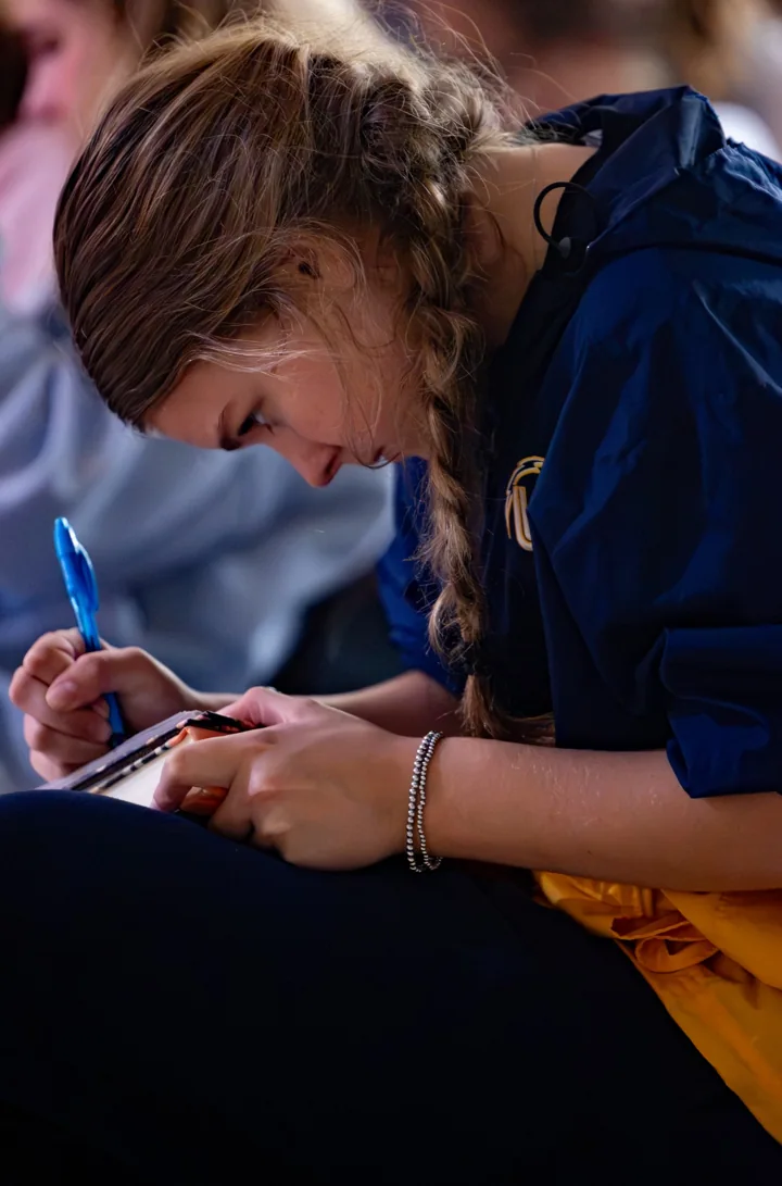 A young woman writes in a notebook while seated at a group gathering.