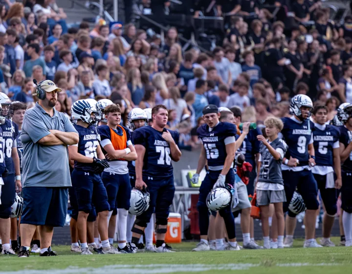 A football team watches the game from the sideline with coaches nearby.