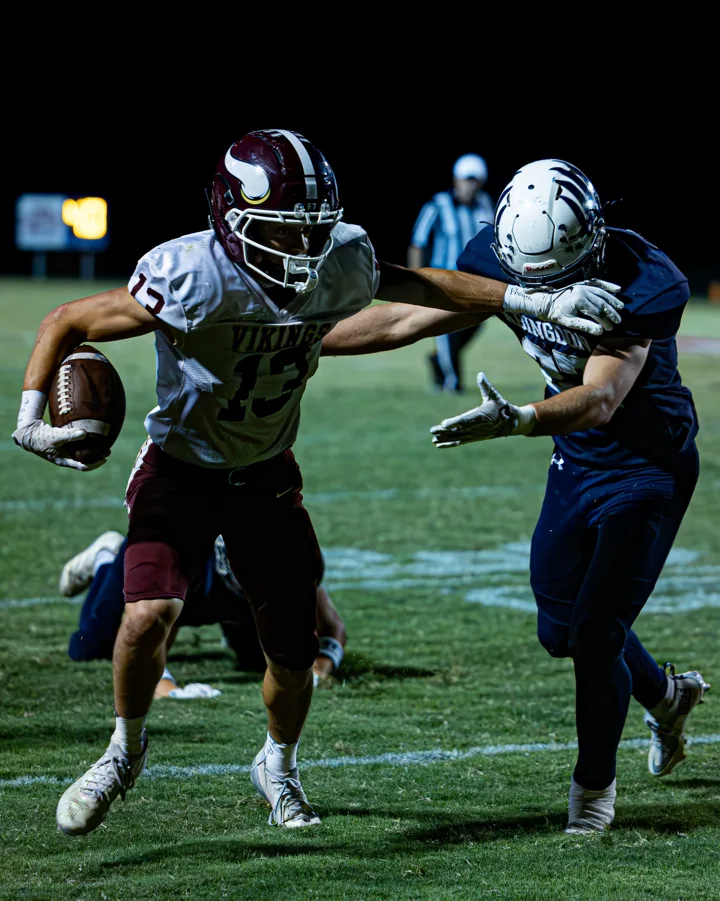 A football ball carrier in white and maroon extends an arm toward an approaching defender.