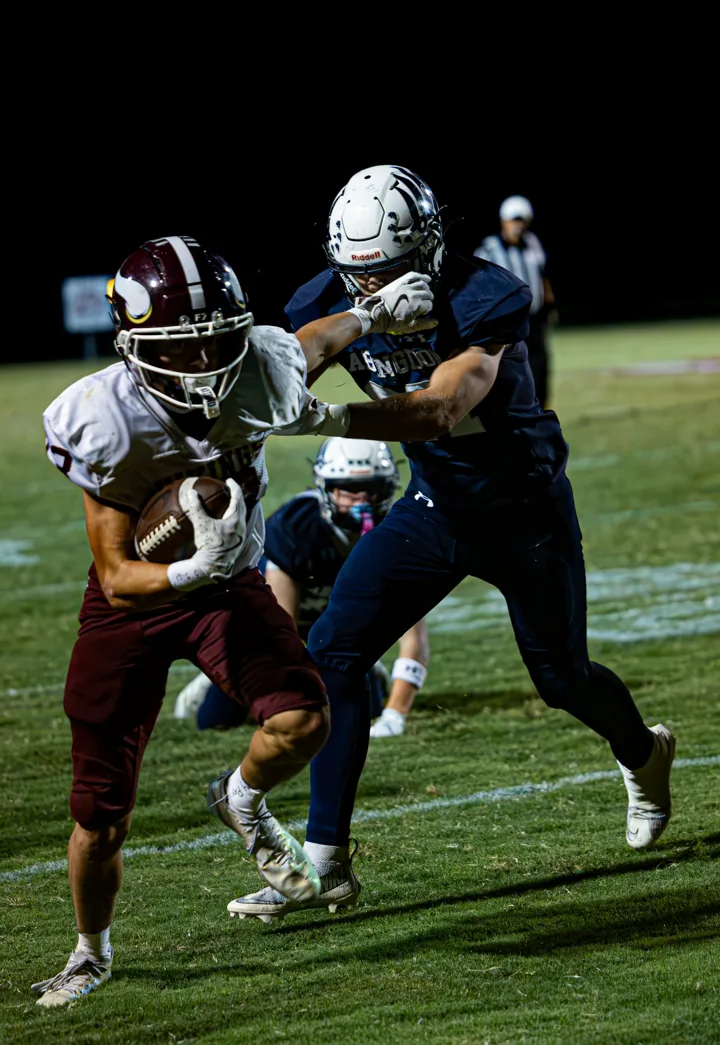 A football ball carrier keeps moving as a navy-clad defender reaches from behind.