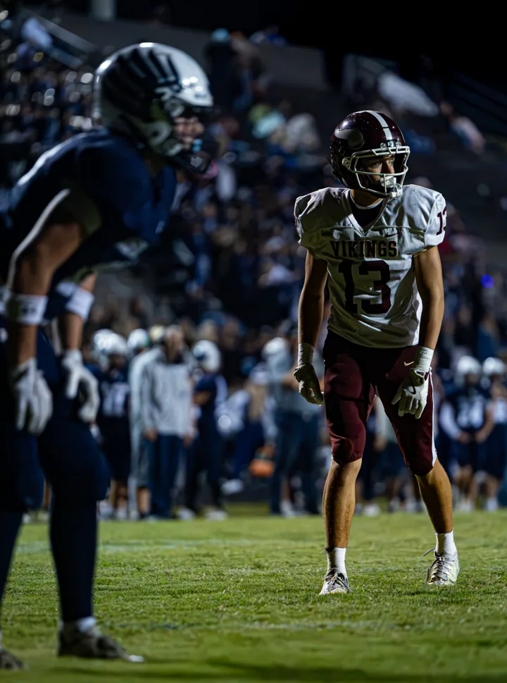 A football player in white and maroon watches opponents across the line under stadium lights.
