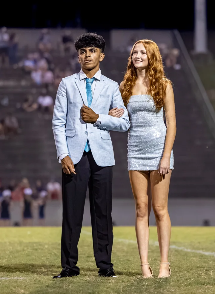 Two people stand arm in arm in formal clothes on the football field.