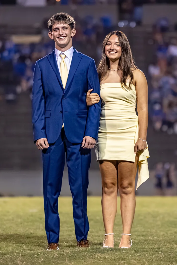 Two smiling people pose together in formal clothes on the football field.