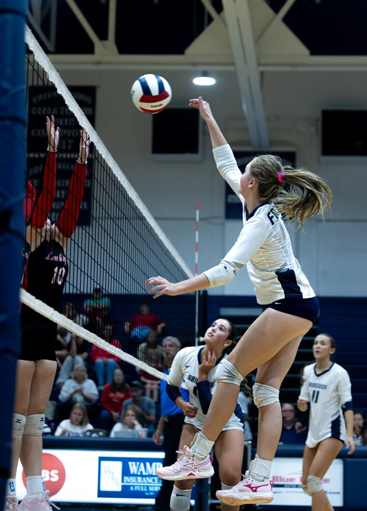 A volleyball player in white jumps to strike the ball above the net.