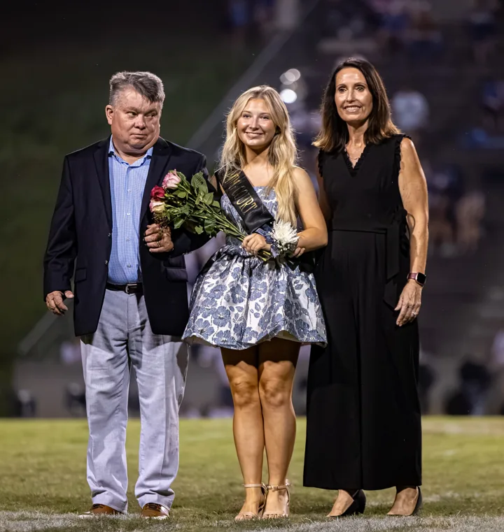 A young woman holding flowers poses between two adults on the football field.
