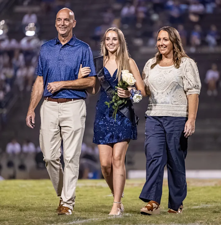 A young woman holding flowers walks across the field between two adults.