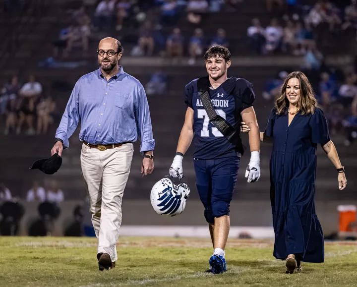 A football player carrying his helmet walks between two adults on the field.