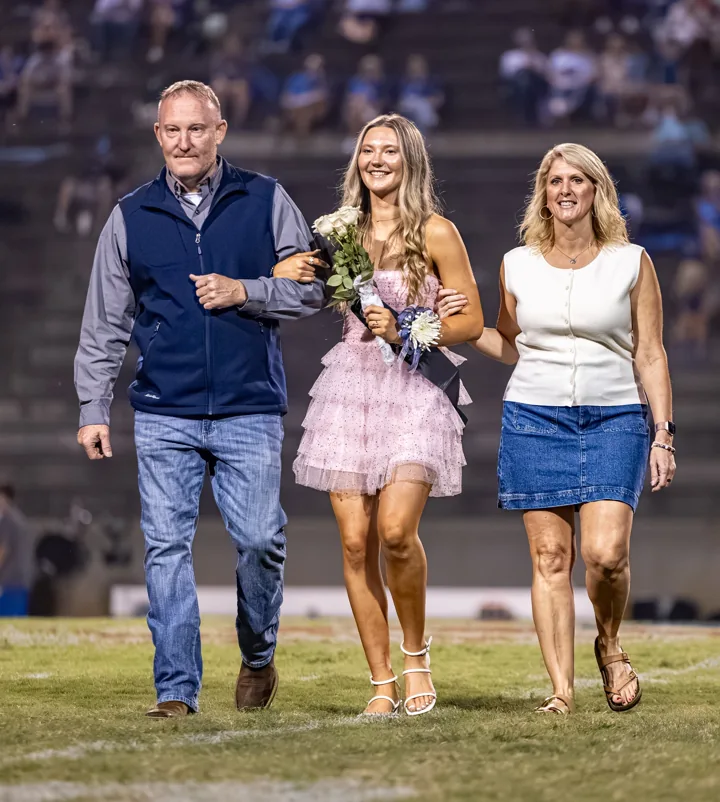 A young woman holding flowers walks arm in arm with two adults.