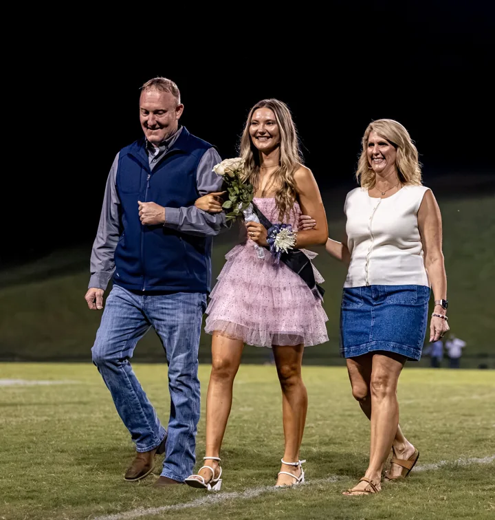 Three smiling people walk together during a football field recognition ceremony.