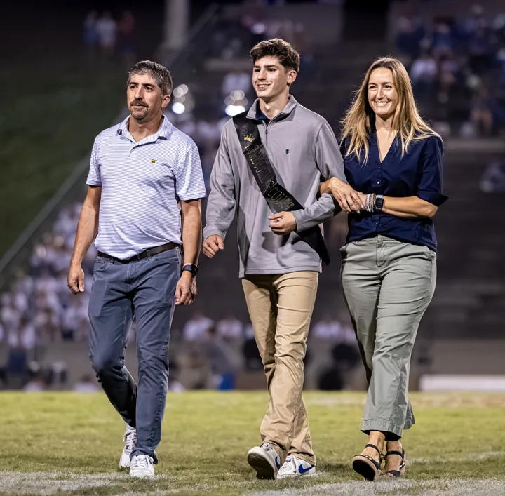 A young man in a vest walks across the field between two adults.