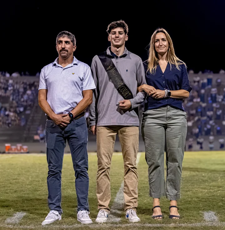 A young man in formal clothes poses with two adults beneath stadium lights.