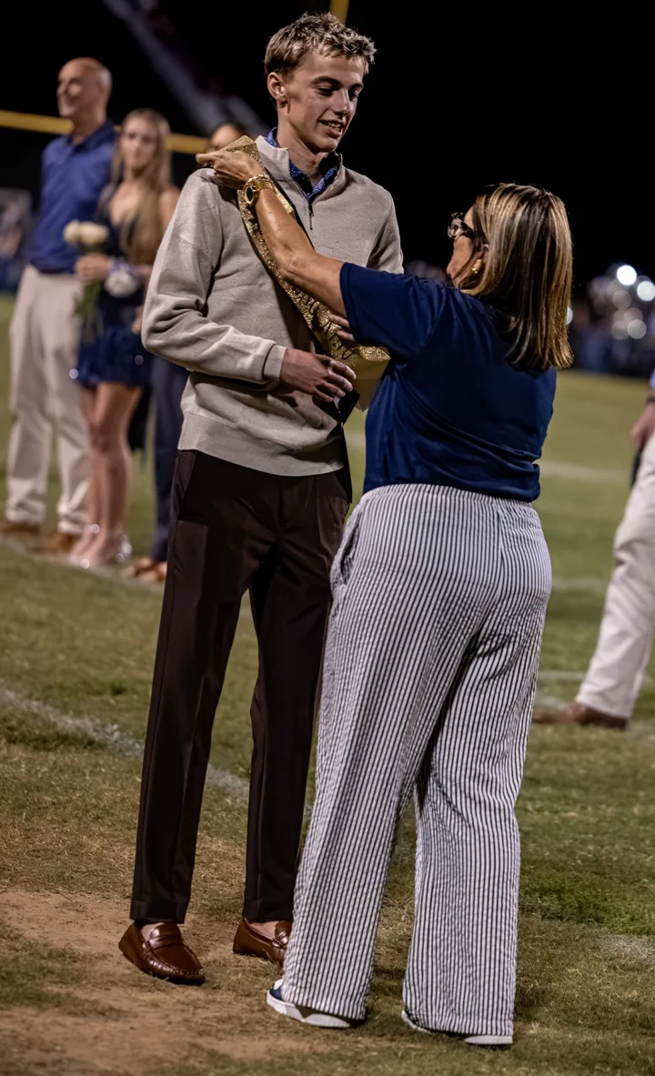 A young man stands while a presenter adjusts a sash during a ceremony on the field.