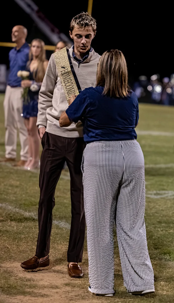 A presenter speaks with a young man wearing a sash on the football field.