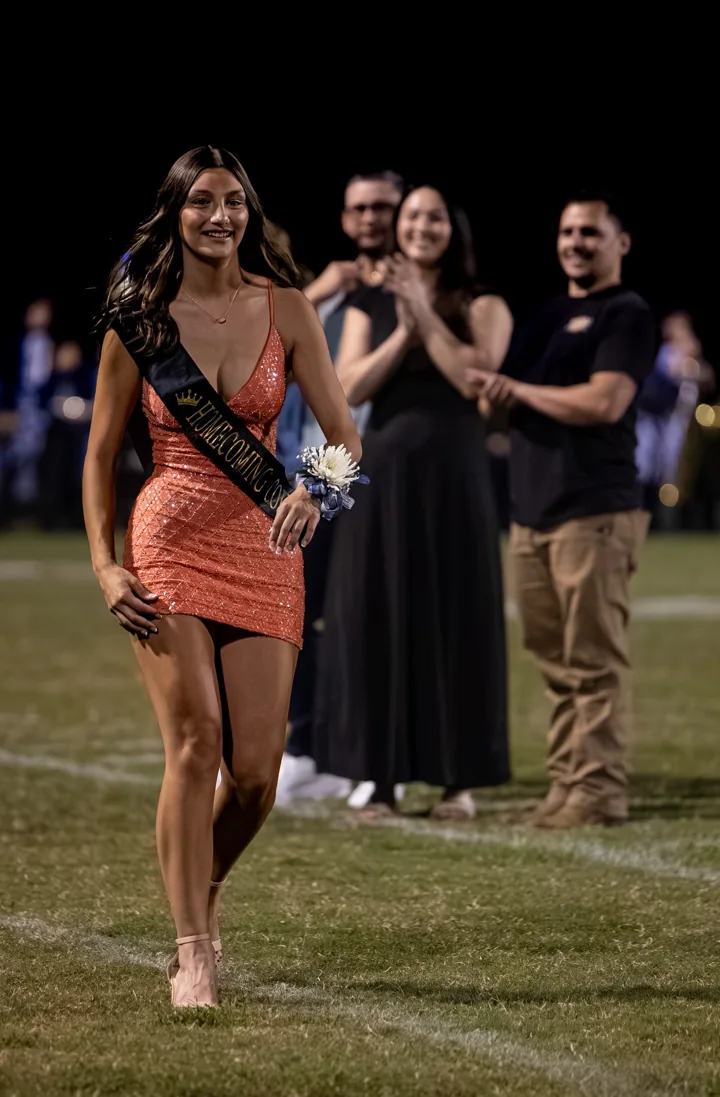 A smiling young woman steps forward with flowers while people applaud behind her.