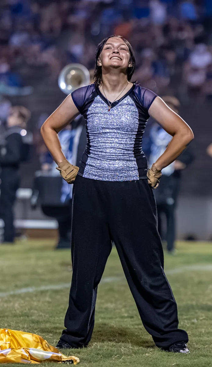 A smiling color guard performer pauses with a gold flag resting on the field.