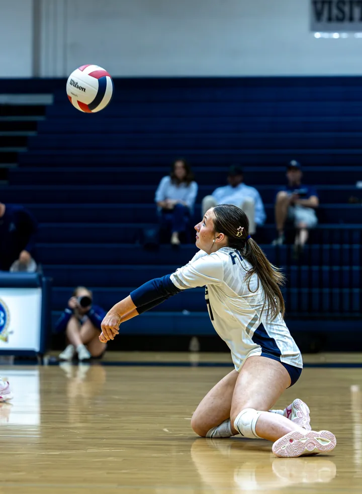 A volleyball player in white reaches from her knees to pass the ball.