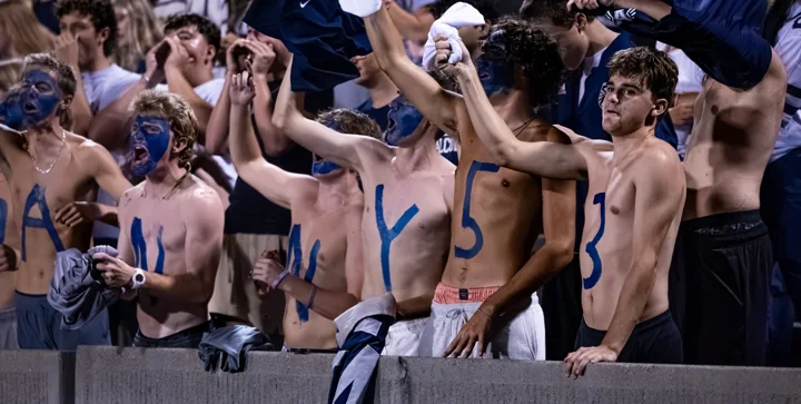 Students in the football grandstand raise their arms and cheer.