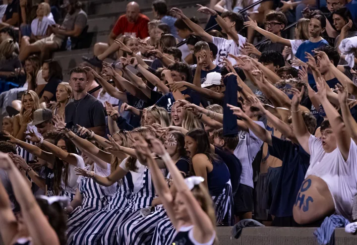 A large student section reaches and cheers together in the football grandstand.