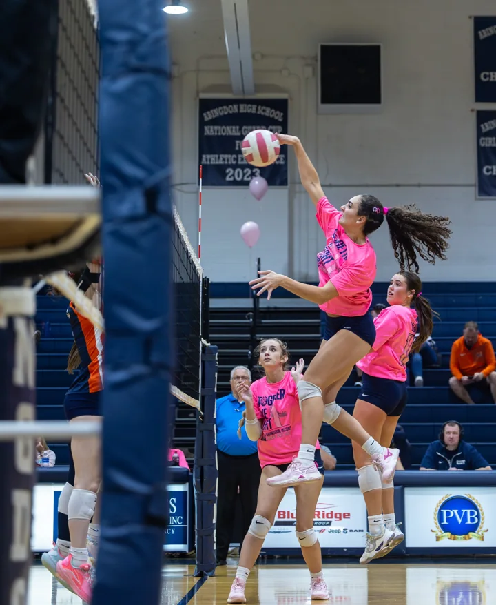 A volleyball player in a bright pink shirt jumps to hit the ball above the net.