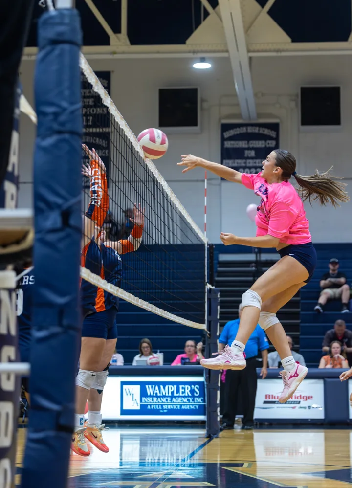 A volleyball player in a bright pink shirt reaches to attack as an opponent prepares to block.