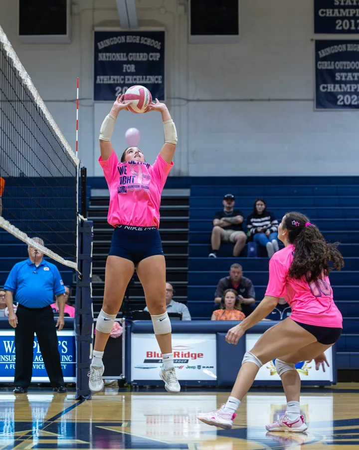 A volleyball player in a bright pink shirt sets the ball with both hands above her head.