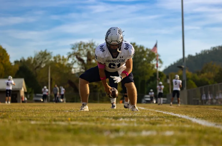 A helmeted football player crouches on the field before the game, framed by blue sky and autumn trees.