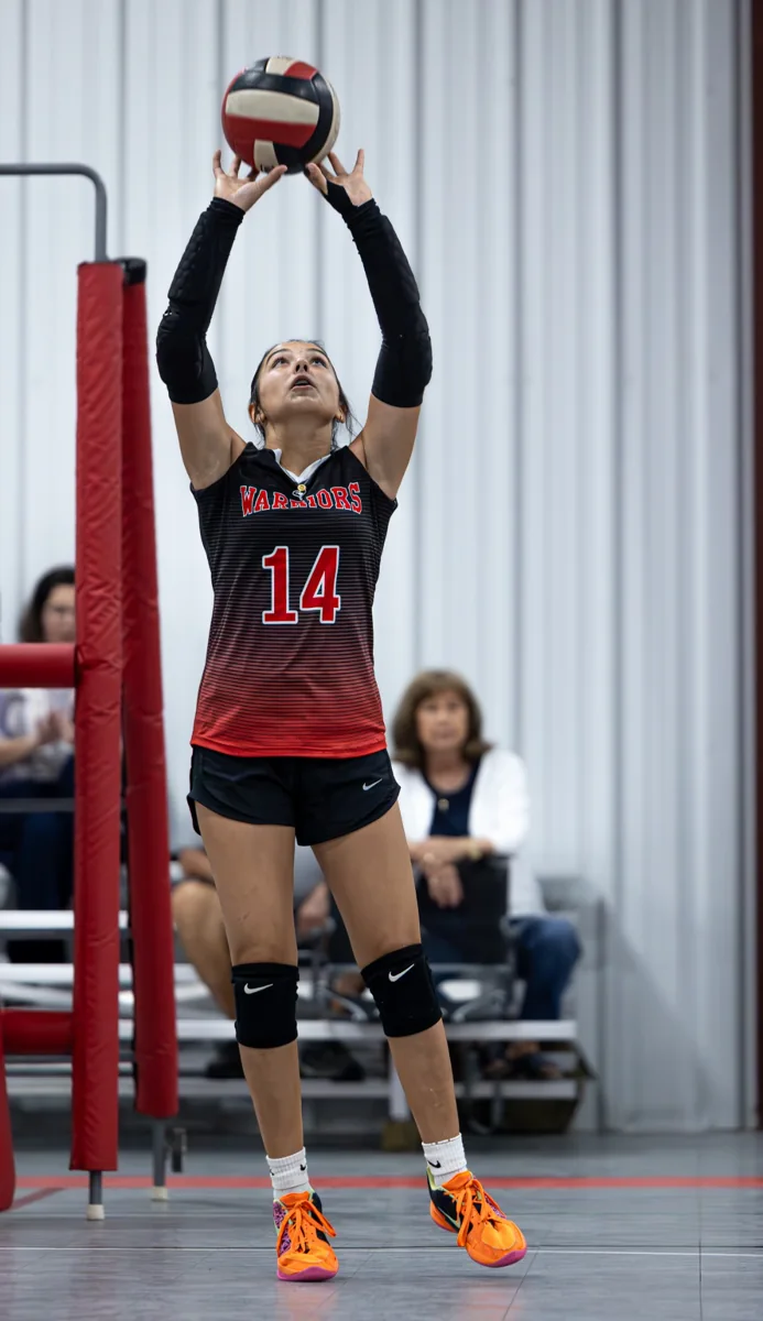 A volleyball player in a red number 14 uniform sets the ball above her head.