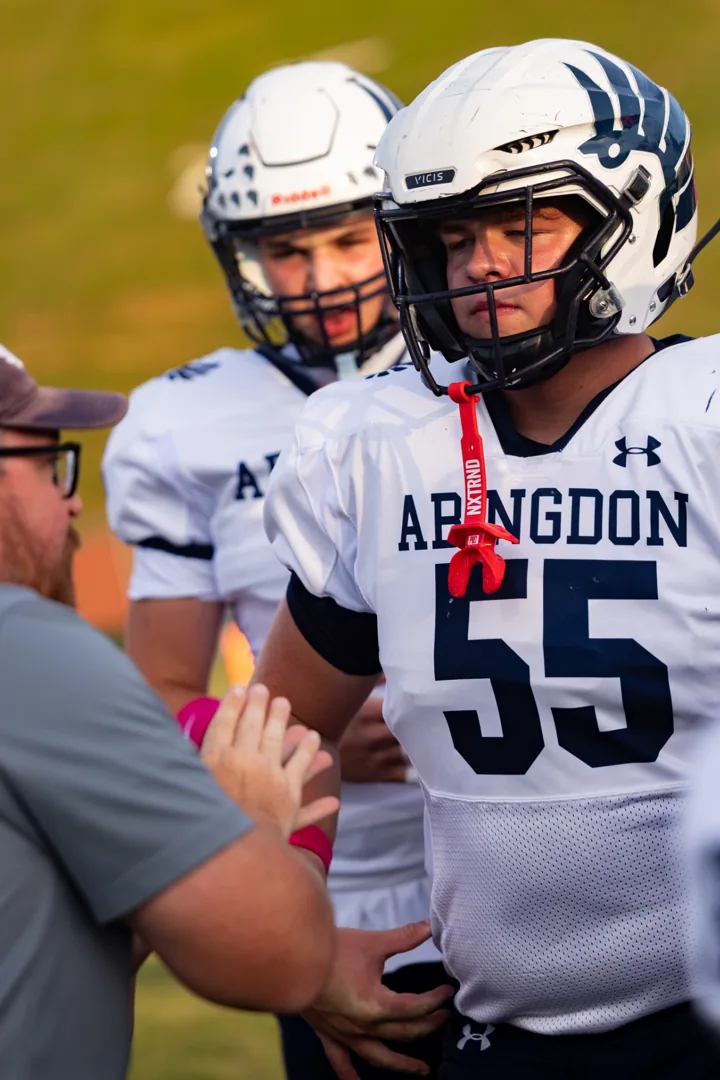 A football player wearing number 55 listens on the sideline with teammates behind him.