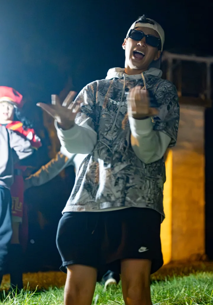 A young man in sunglasses smiles and gestures outdoors under evening event lights.