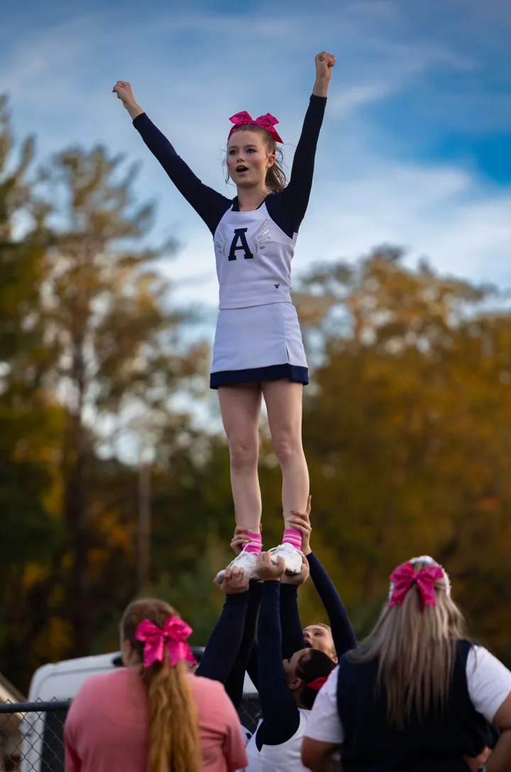 A cheerleader stands elevated above her teammates with both arms raised.
