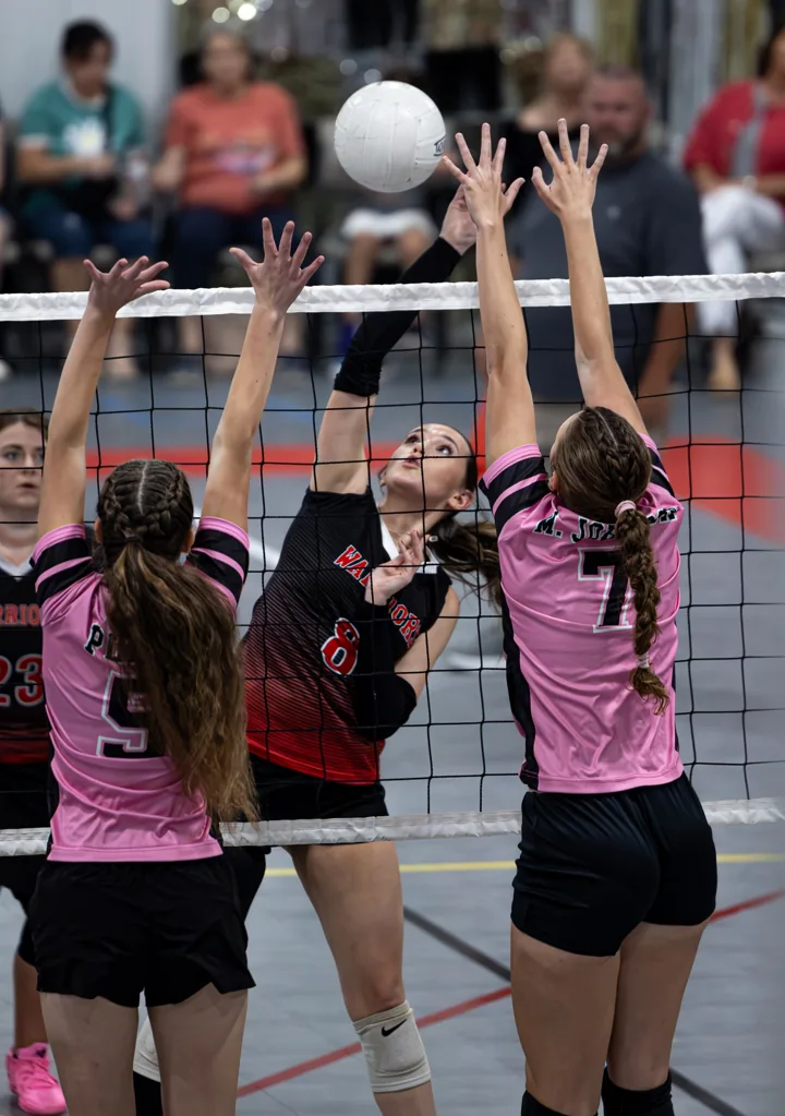 A volleyball player in red attacks the ball against two blockers in pink.