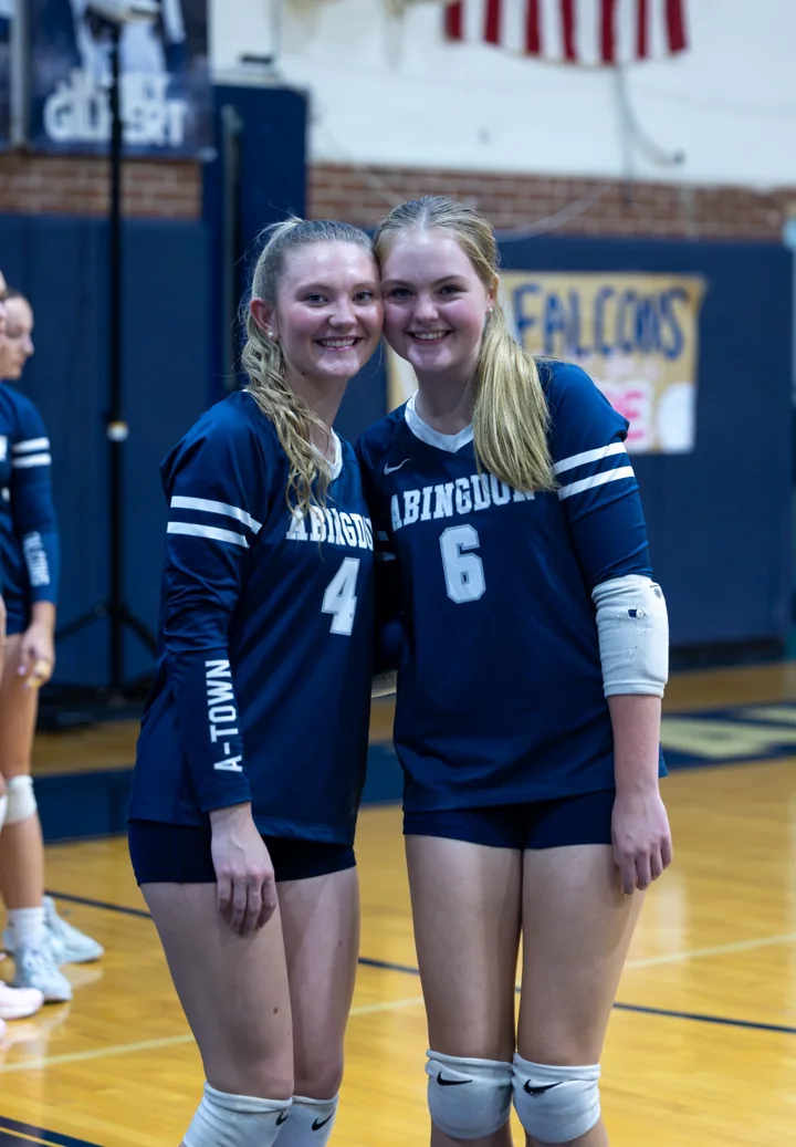 Two volleyball teammates in navy uniforms smile together on the court.