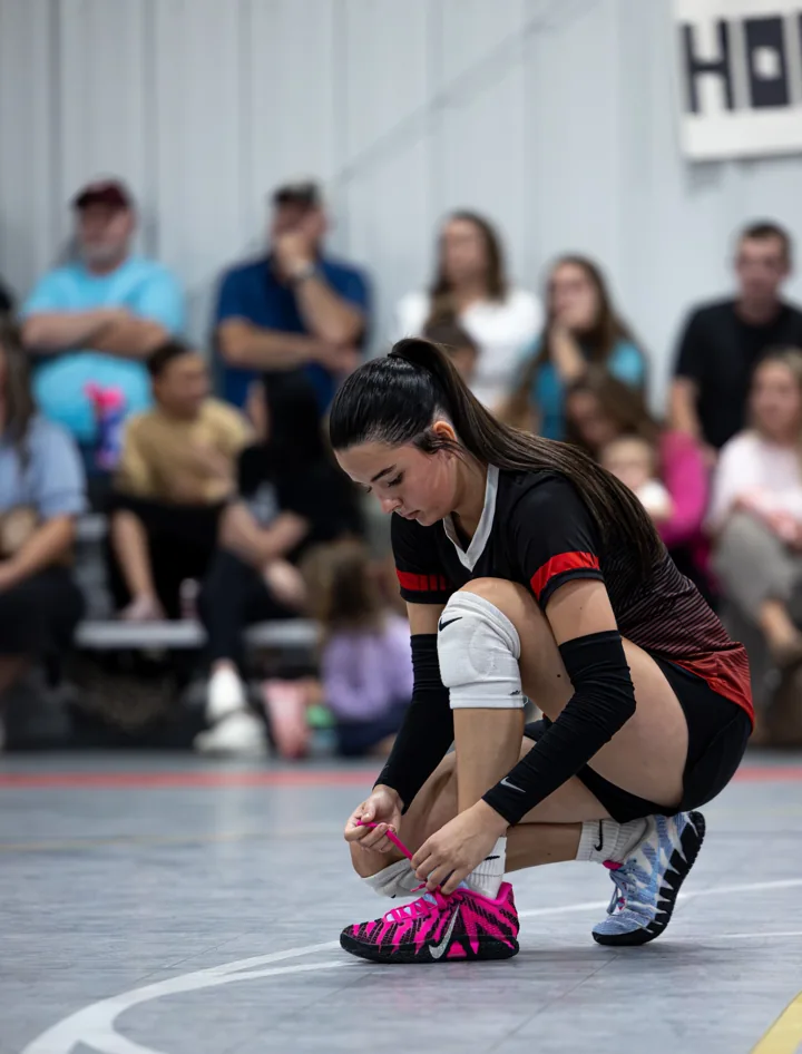 A volleyball player in red and black bends down to tie a bright pink shoe.