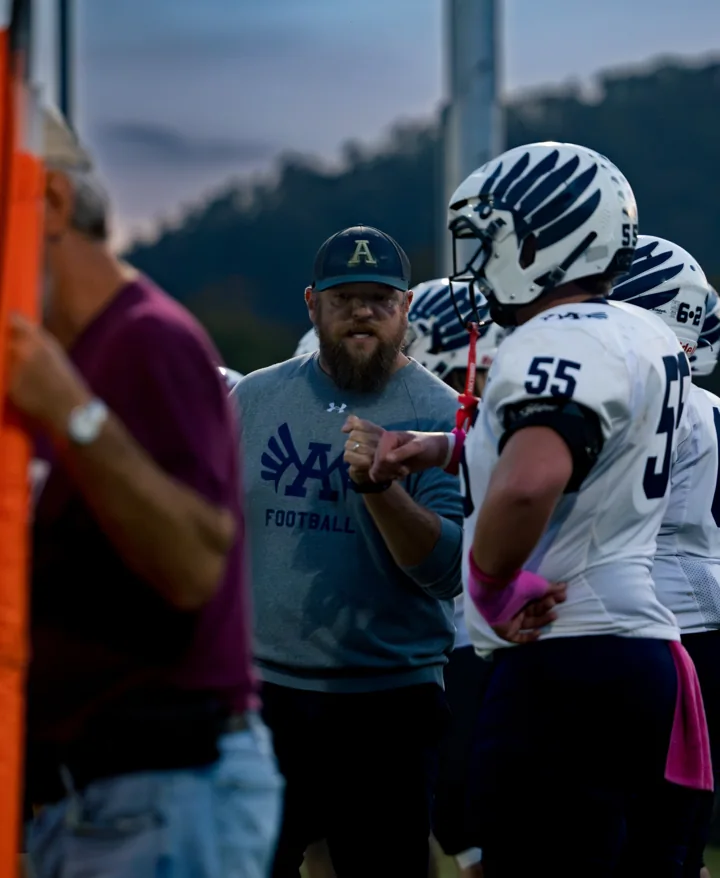 A coach talks with a helmeted football player on the sideline.