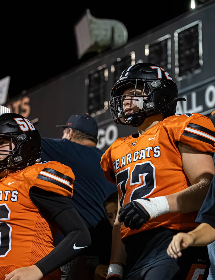 A helmeted football player in an orange uniform stands with teammates under stadium lights.