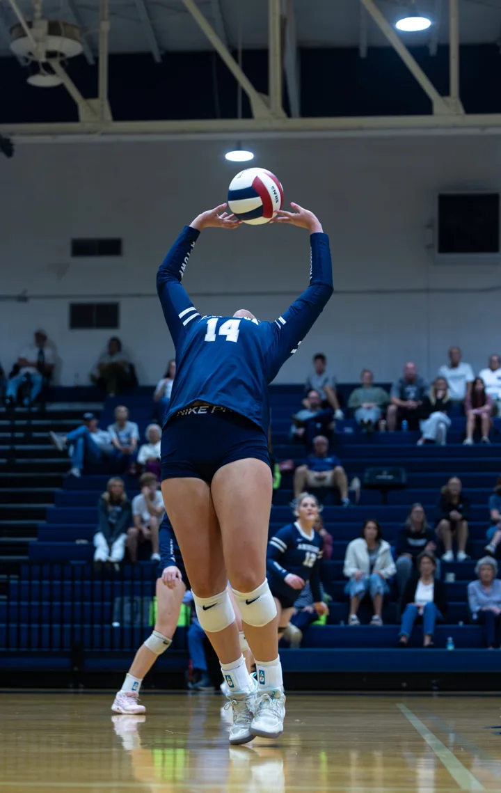 A volleyball player in a navy number 14 uniform jumps to set the ball overhead.