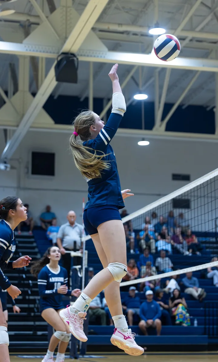A volleyball player in navy stretches upward to attack the ball above the net.