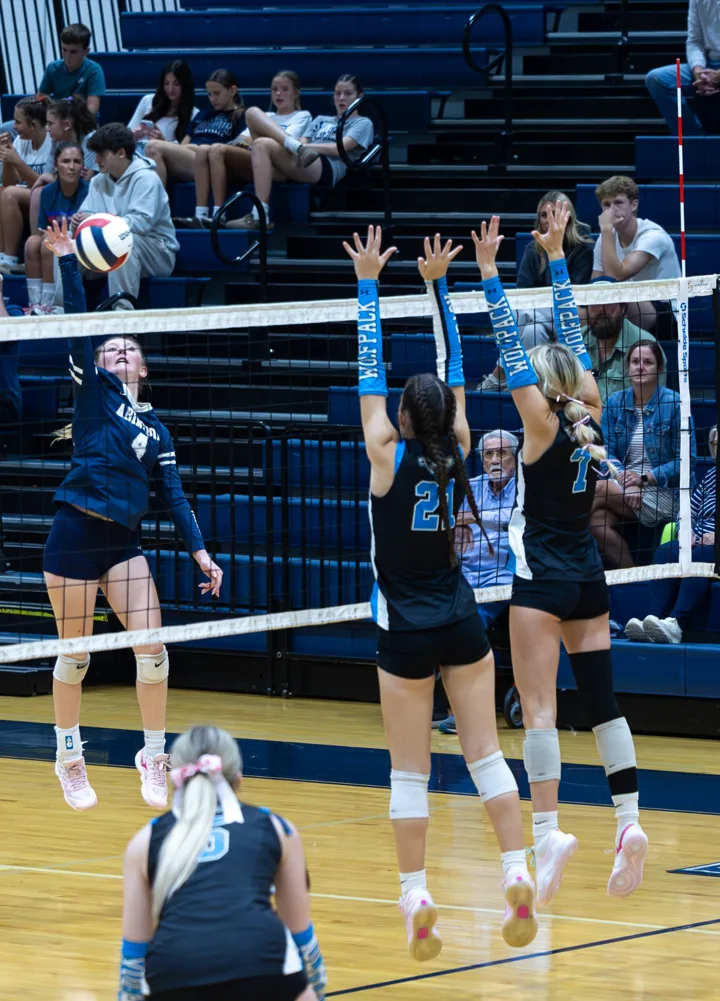 A volleyball attacker in navy faces two blockers at the net.