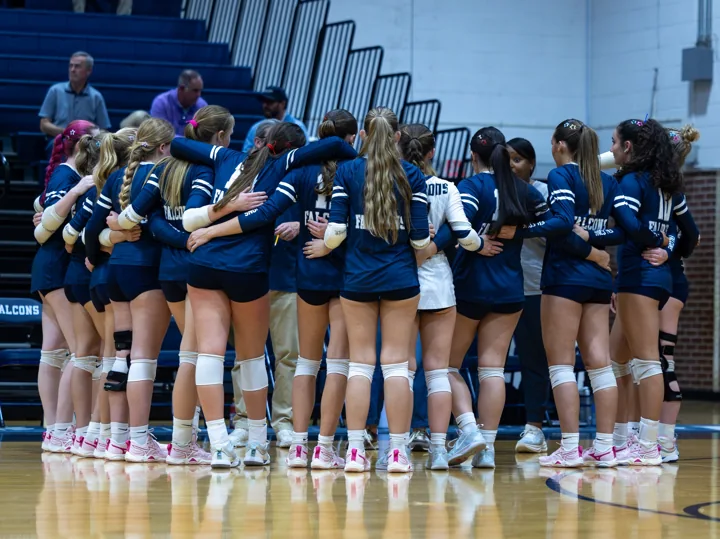 A volleyball team gathers shoulder to shoulder in a close court huddle.